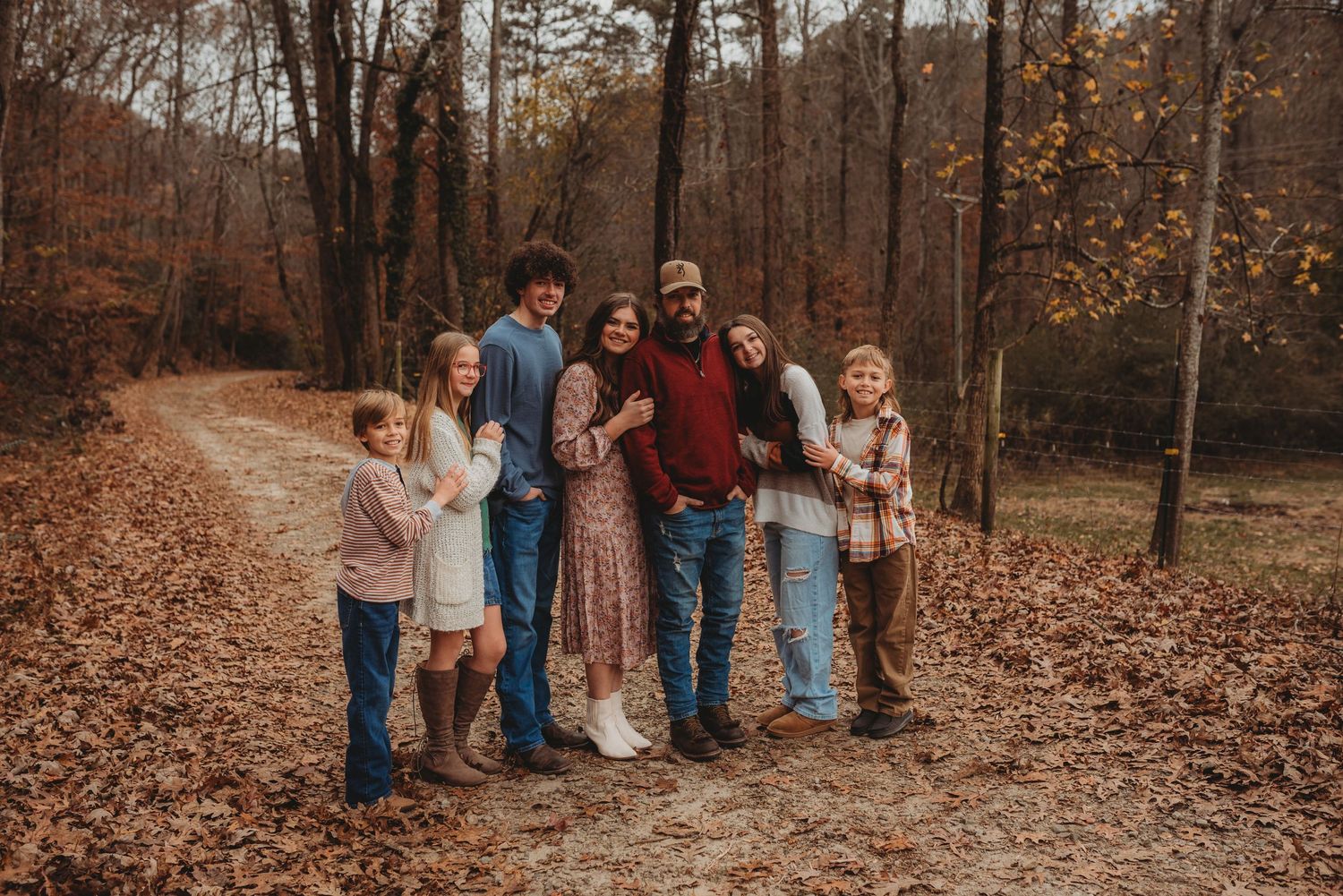 Large family poses together on a leaf-covered autumn trail surrounded by fall foliage.