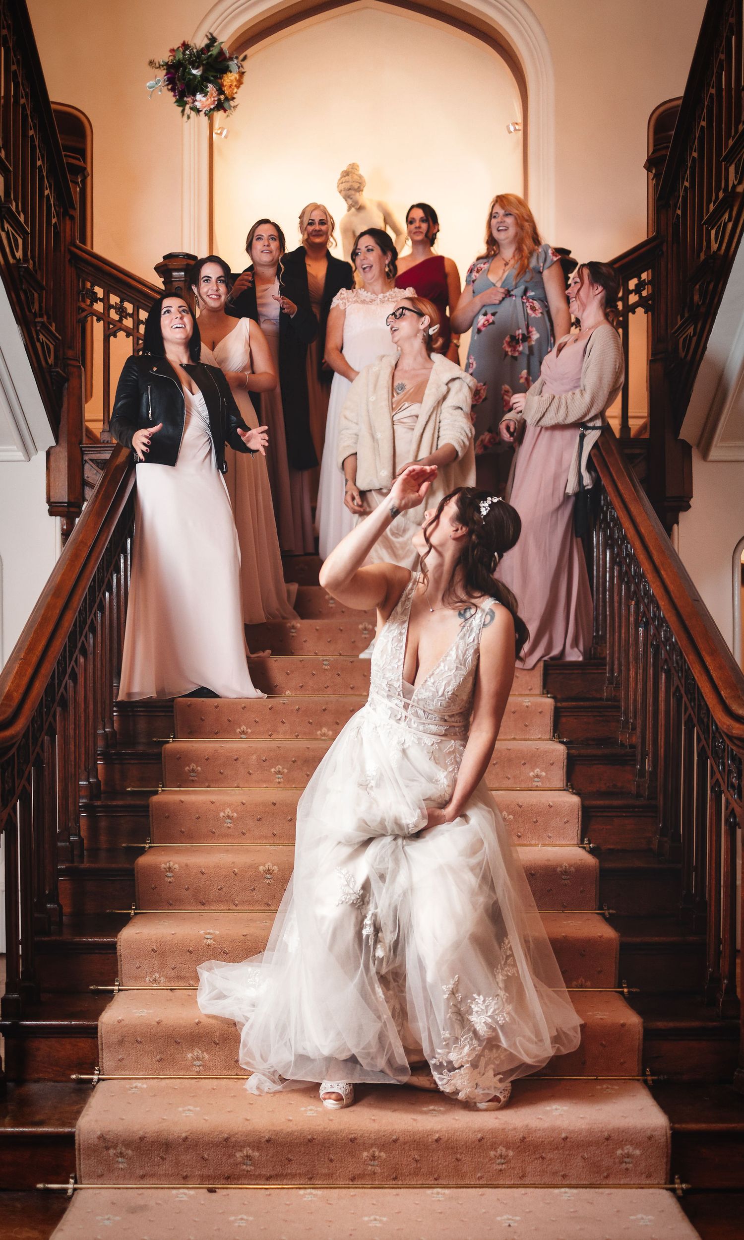 Bride on a grand wooden staircase with wedding party gathered above, elegant venue wedding photography Southampton