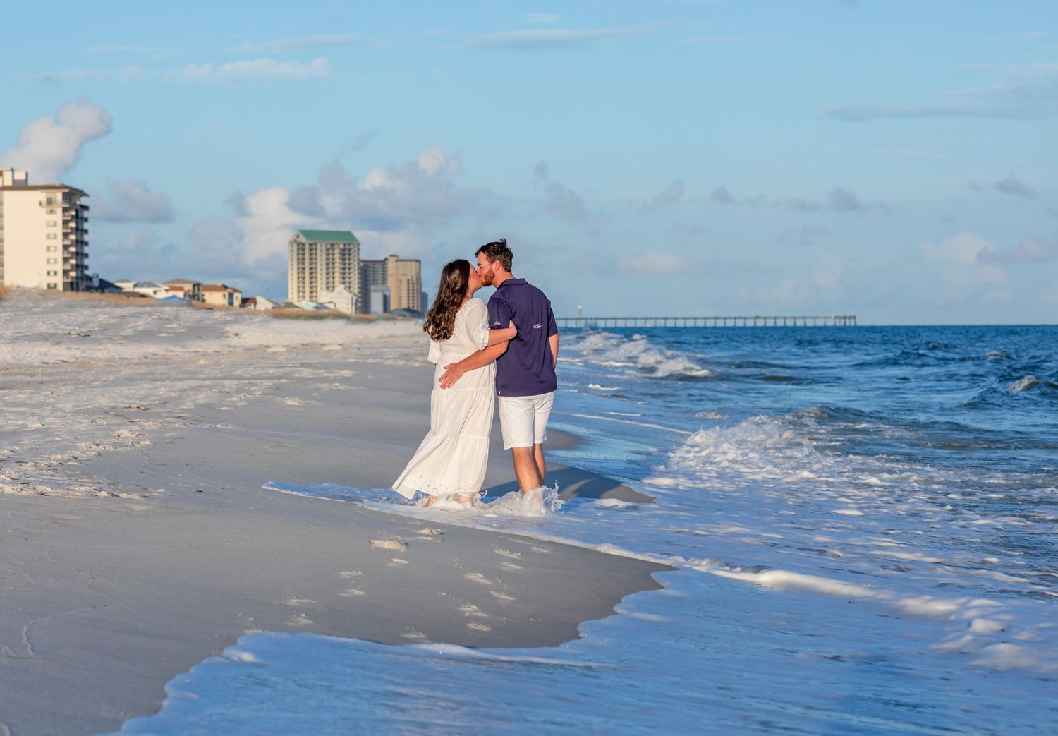 Romantic beach scene with couple embracing on sandy shore at sunset with pier in distance.