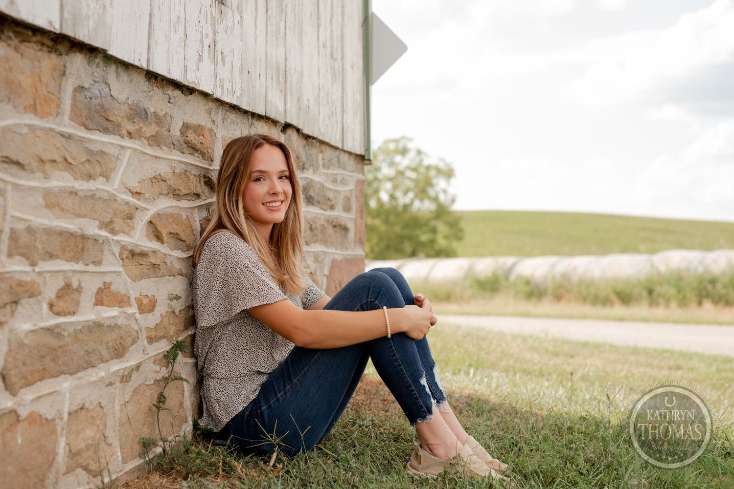 A person in casual attire sits against a stone and wood barn wall in a rural setting during golden hour.