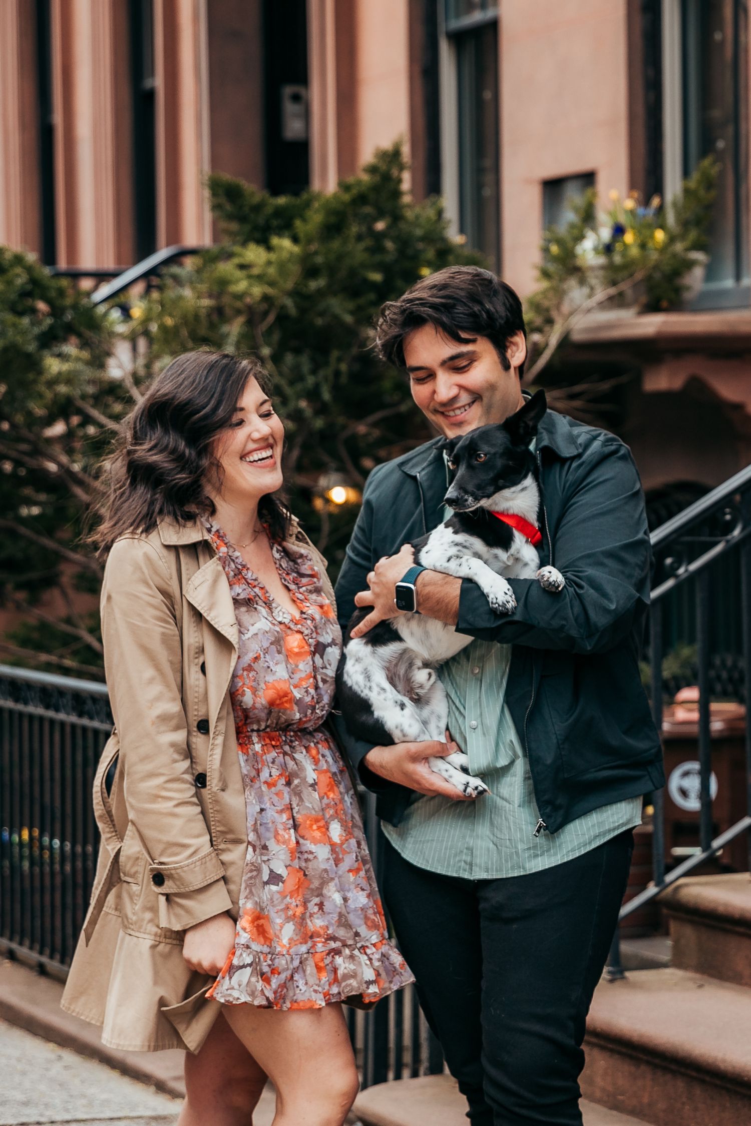 Happy couple laughing while man holds small black and white dog on city brownstone street.