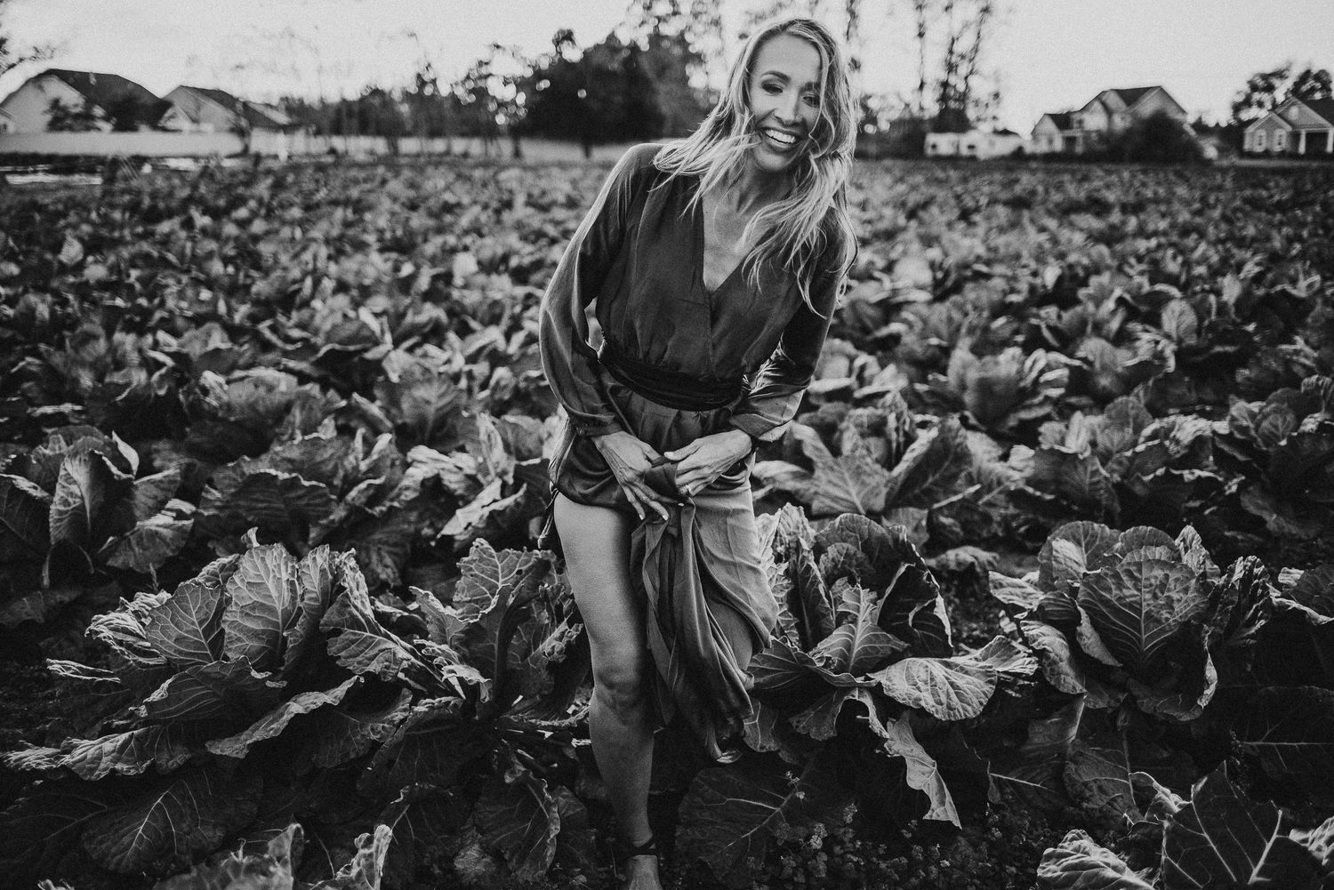 Woman in dark dress poses playfully in large cabbage field on farm.