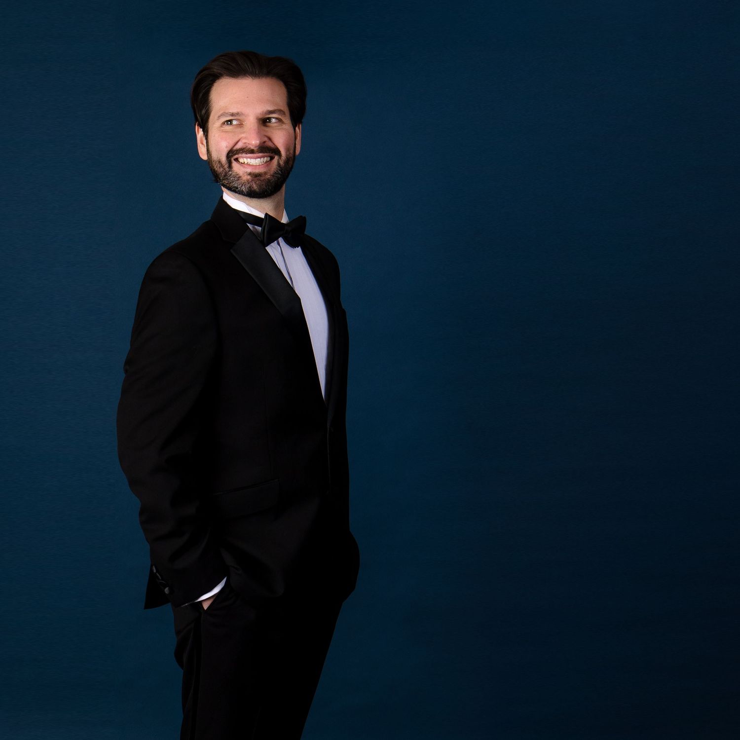 Formal portrait of person in tuxedo against deep blue background.