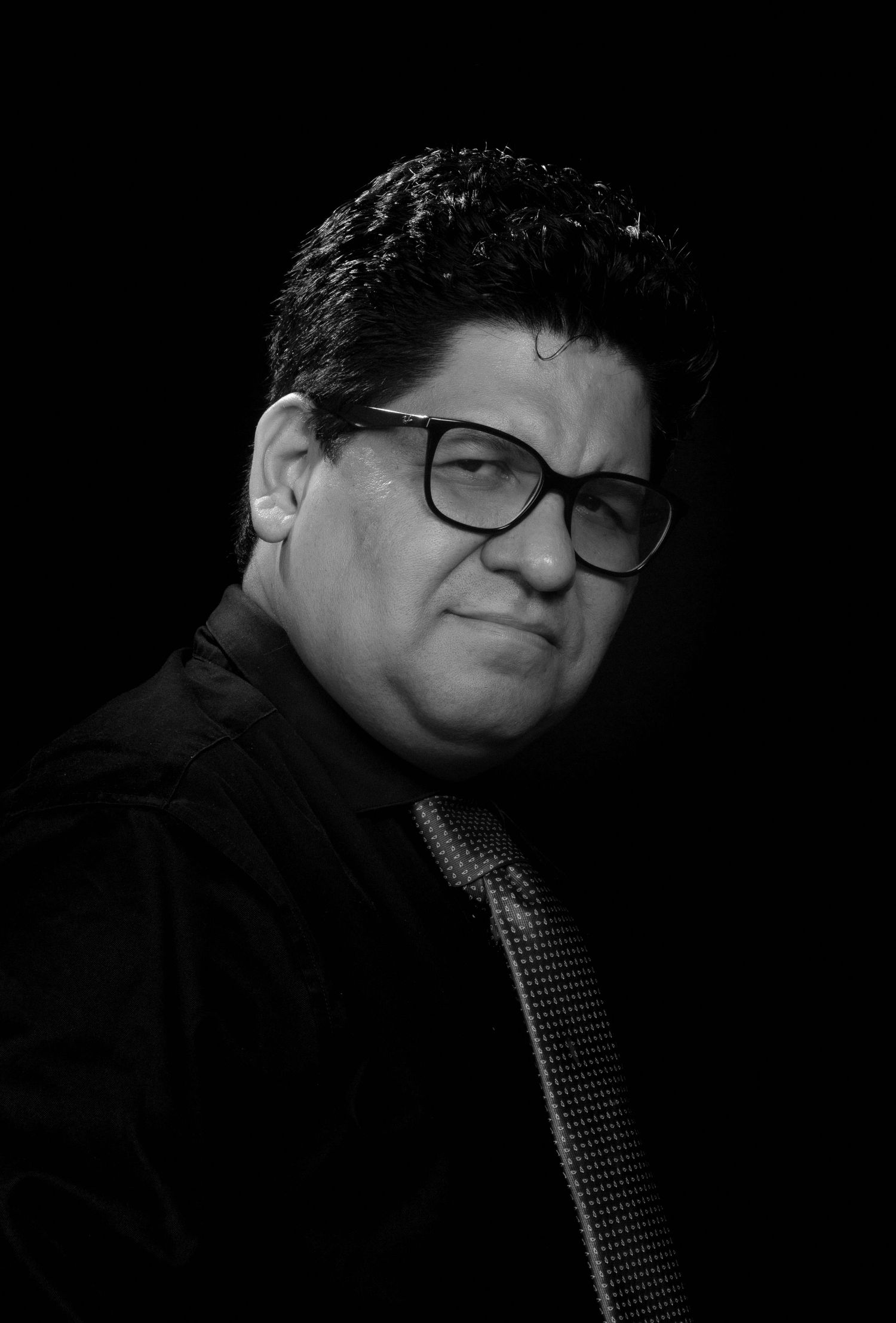 Black and white portrait in dramatic lighting of someone wearing glasses and a suit against a dark background.
