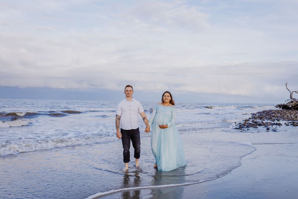 A couple stands at the ocean's edge wearing light colors during their maternity photoshoot.