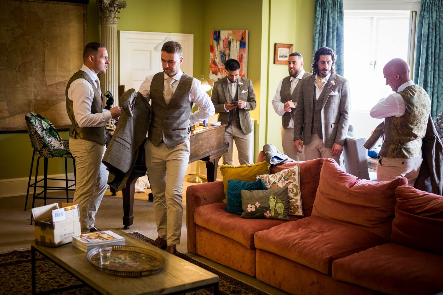 Wedding party members in vests and formal attire gather in a living room with a brown couch.