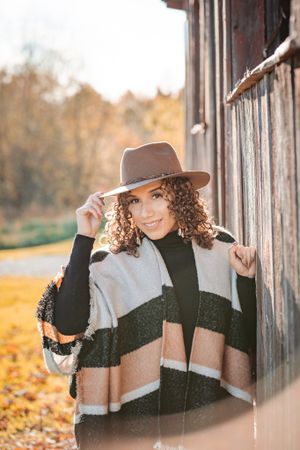 Woman in cozy striped sweater and brown hat poses near rustic wooden wall with autumn foliage in background.