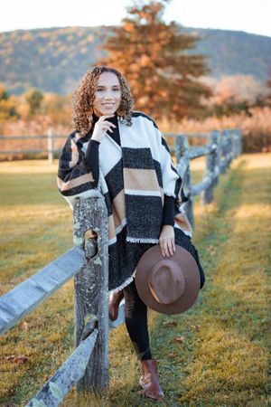 Person in black and white plaid poncho and brown hat poses against rustic wooden fence at sunset in the Hudson Valley,NY.