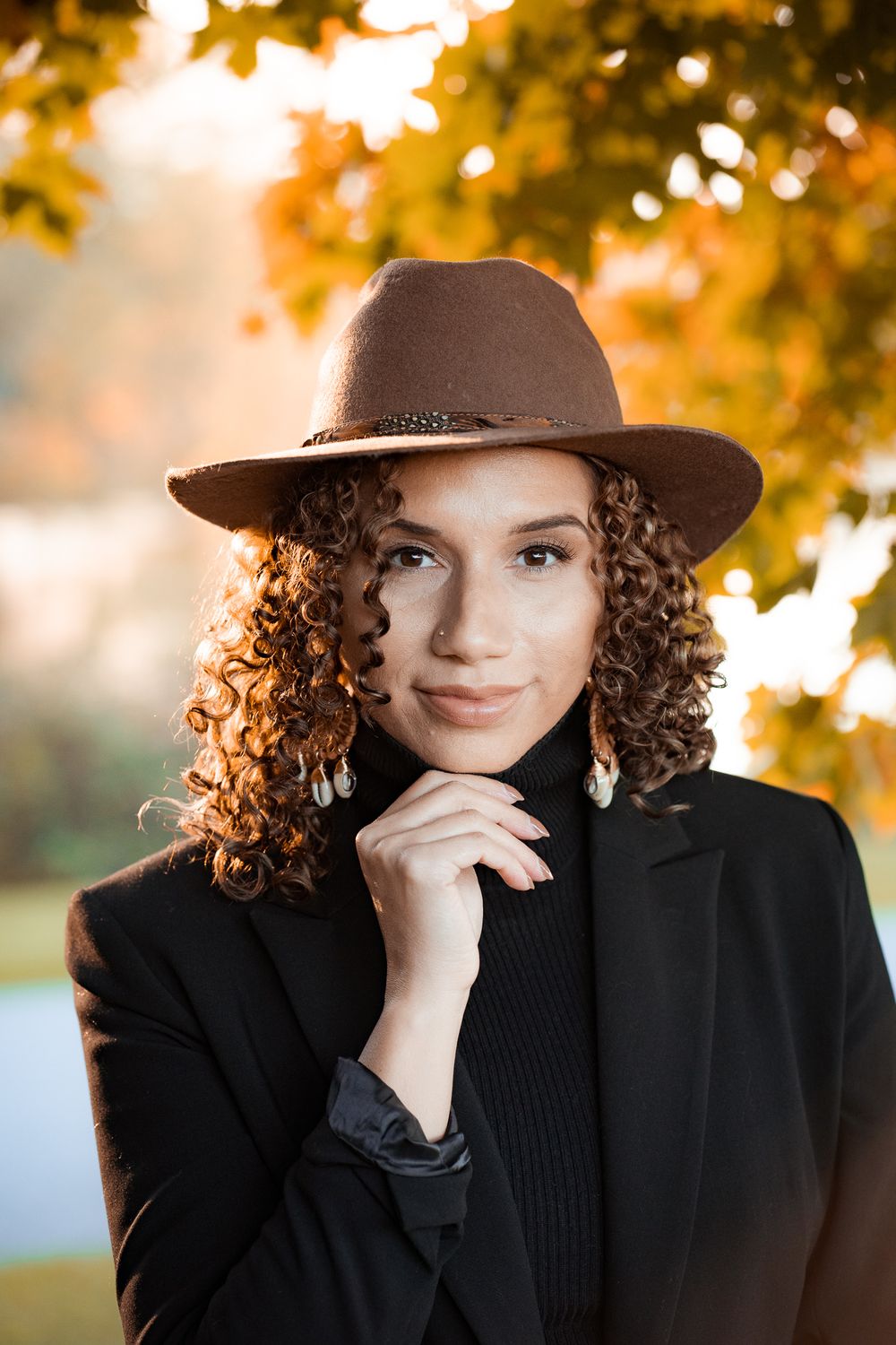 Portrait of a person wearing a brown fedora hat and black blazer against autumn foliage with golden sunlight in background.