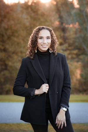 Professional portrait of person in black blazer with curly hair against autumn background.