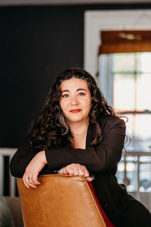 Professional portrait of person in black blazer leaning on brown leather chair.