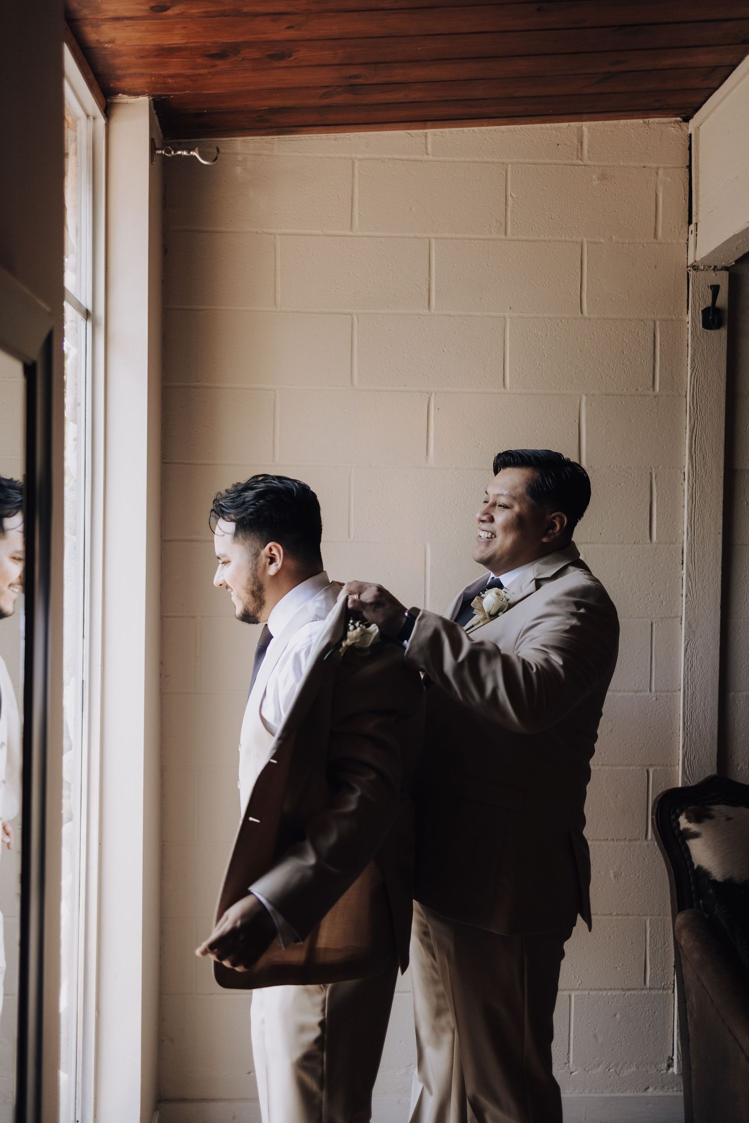 Two people in light colored suits help each other get dressed near a window.
