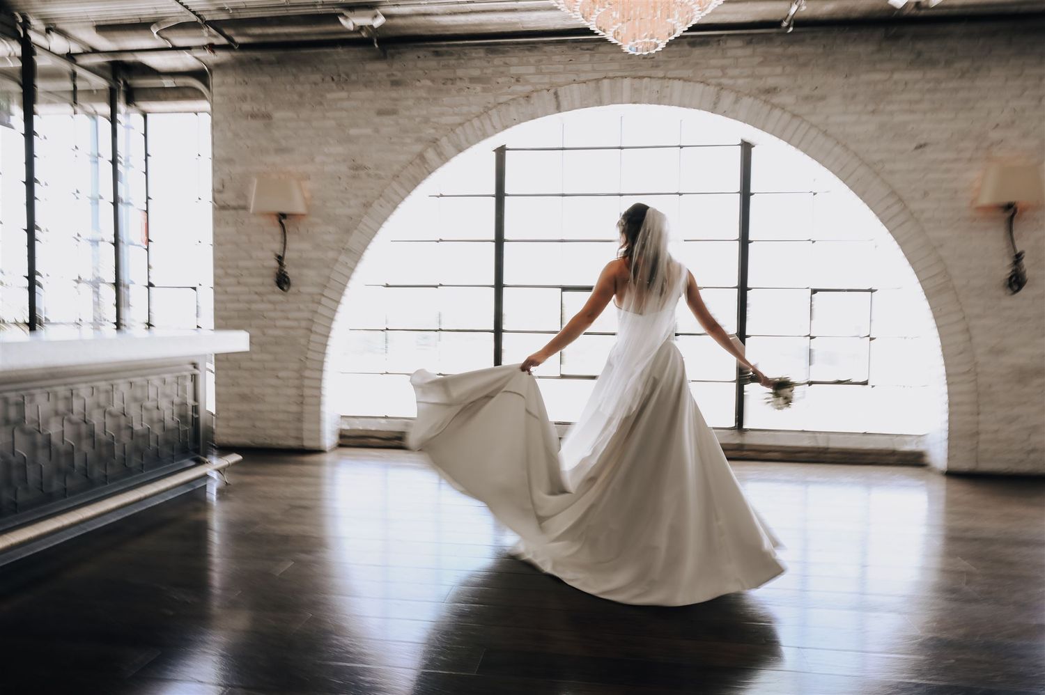 Bride twirling in flowing white dress against industrial brick wall with arched window.