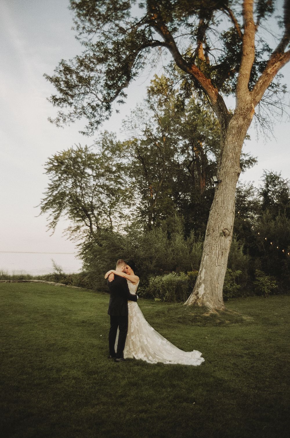 A couple embraces under a large tree during their wedding celebration at sunset.