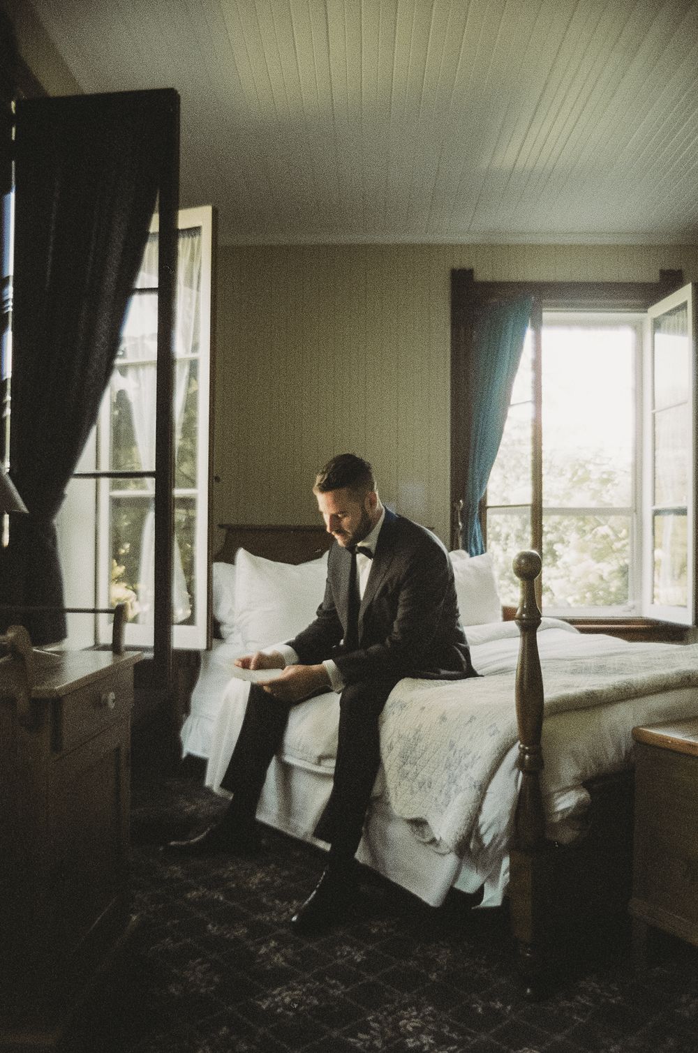 A figure in a dark suit sits on a bed in a sunlit room with white curtains and wooden floors.