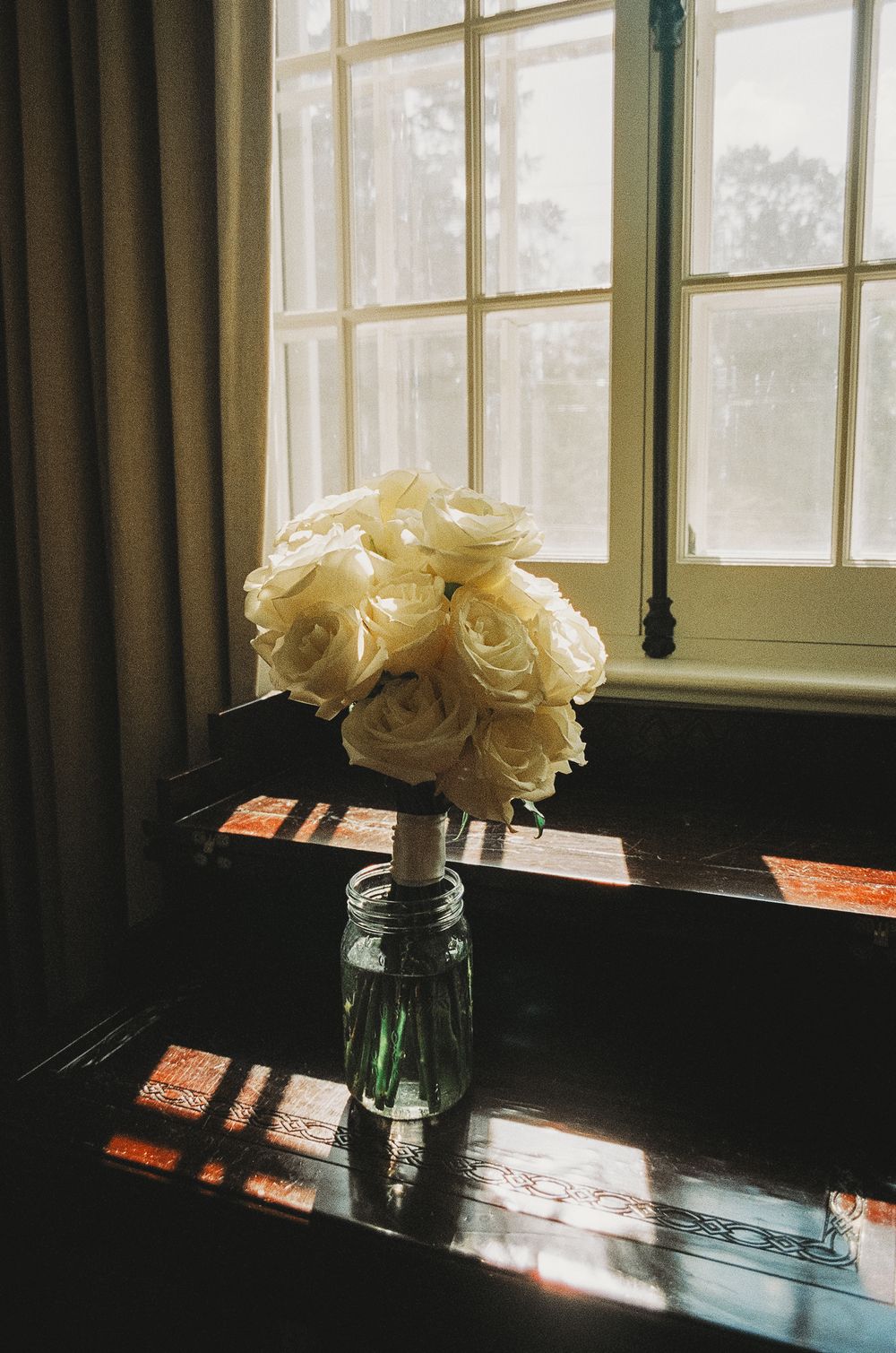White roses in a glass vase catch afternoon sunlight through a paned window.