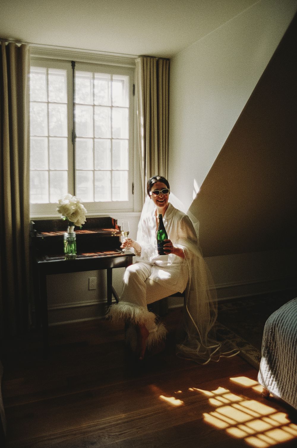 A person in white sits by a window with sunlight streaming in, holding a beverage.