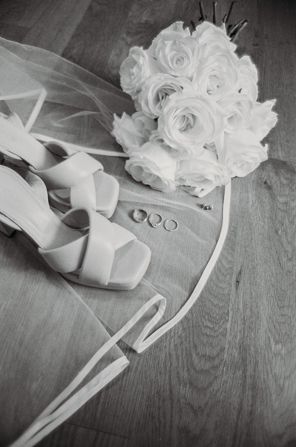 Wedding details including white roses, high heeled shoes, and wedding rings arranged on wooden flooring.