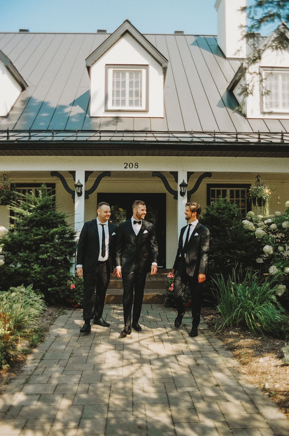 Three men in black suits stand on the front porch of a gray house with dormer windows.