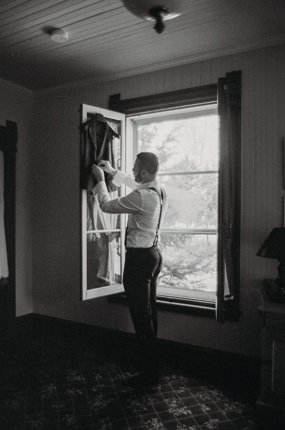 A person in formal attire adjusts clothing by a bright window in a classic interior.