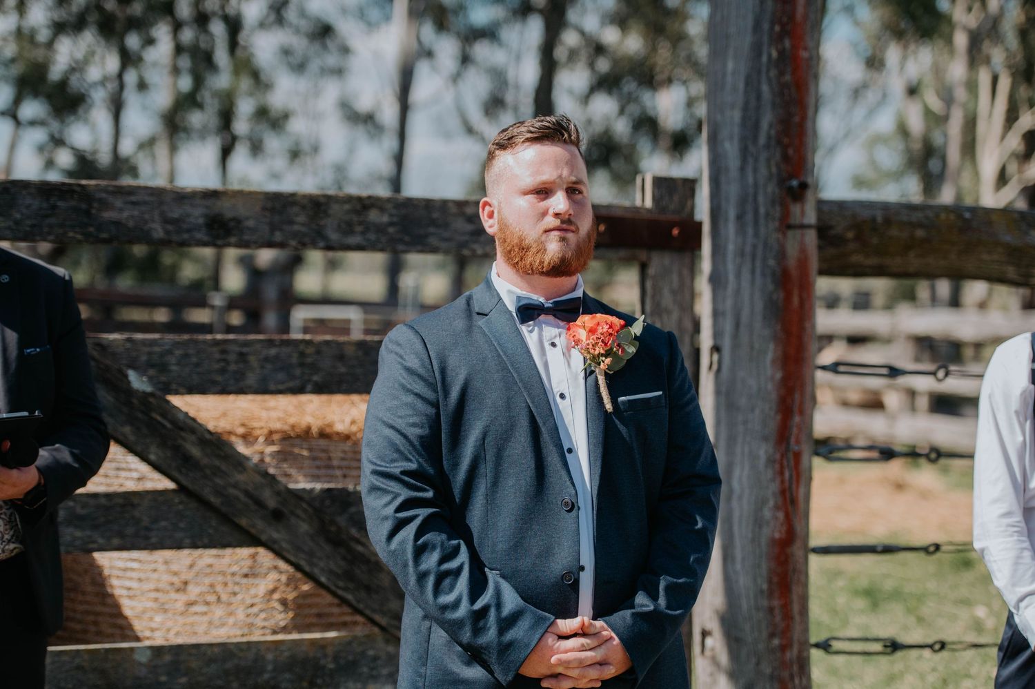 A formally dressed person in a blue suit and orange boutonniere stands in front of a rustic wooden fence.
