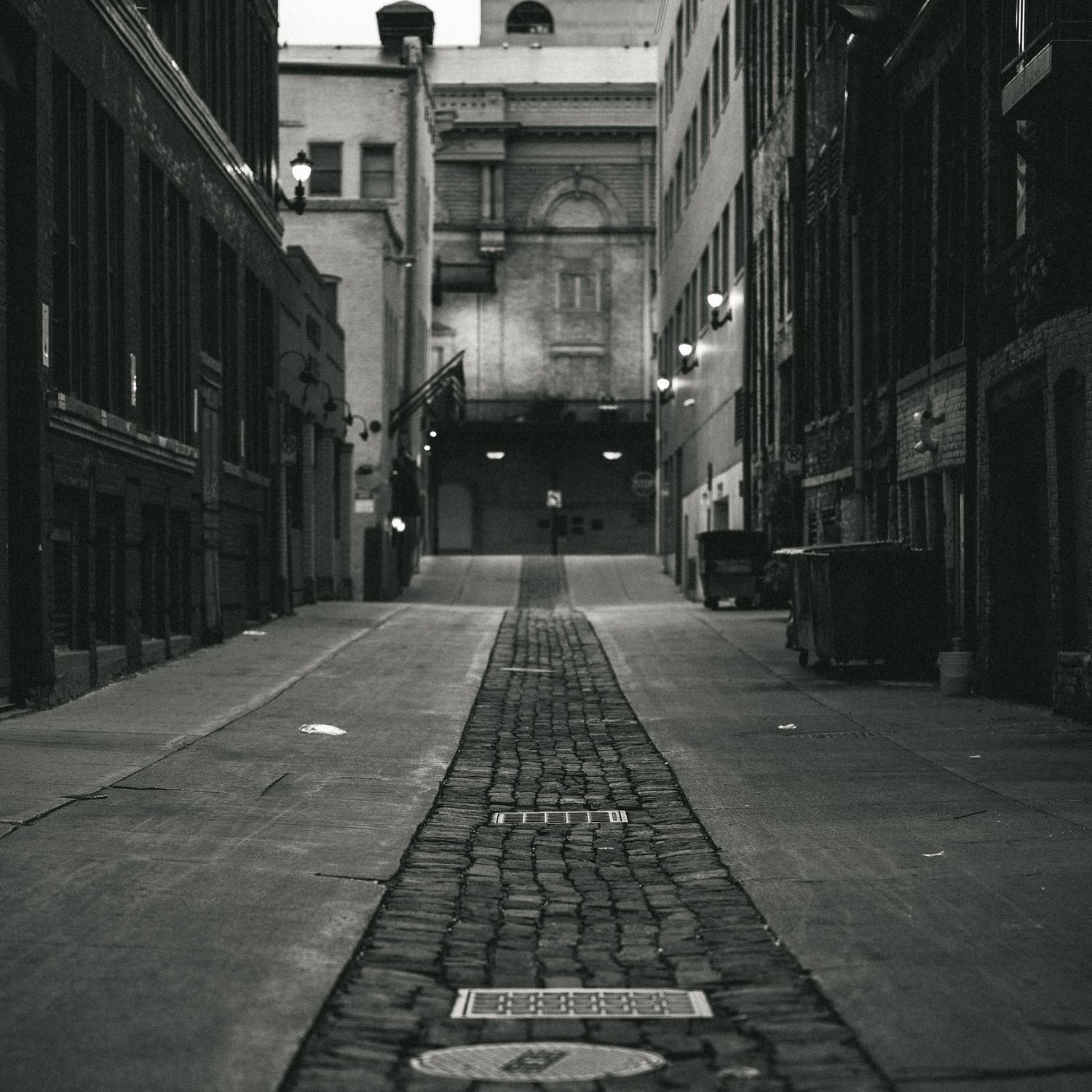 Dark moody alley with cobblestone path leading to illuminated building entrance.
