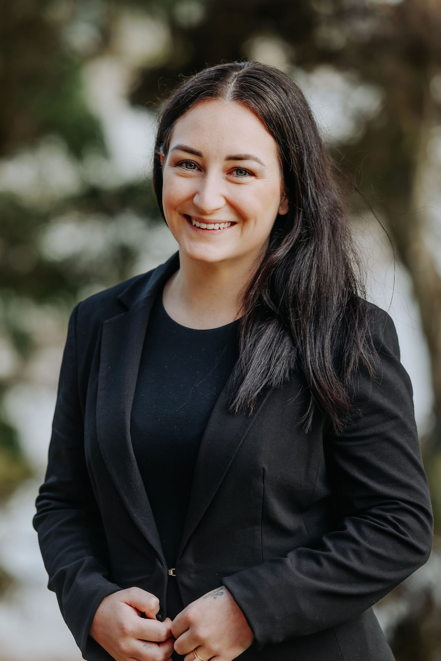 Professional portrait of a person in a black blazer smiling warmly against a blurred outdoor background.