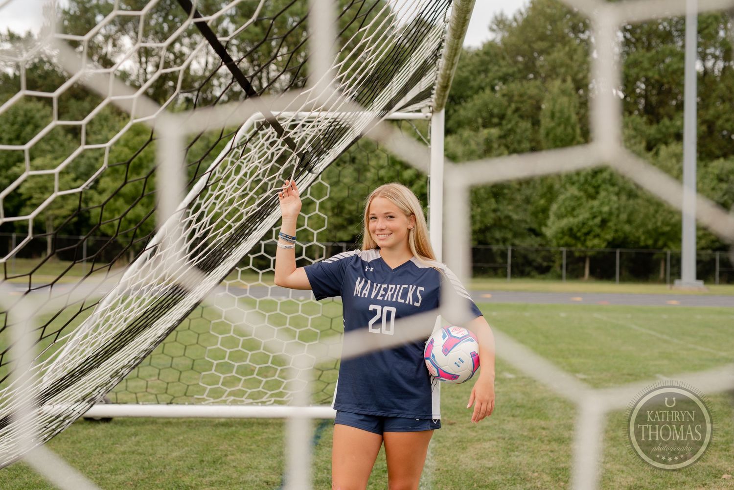 Athlete in Maverick uniform posing with ball through soccer goal netting.