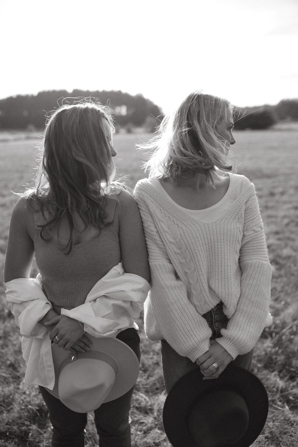 Black and white photo showing the back view of two friends standing close together in a field.