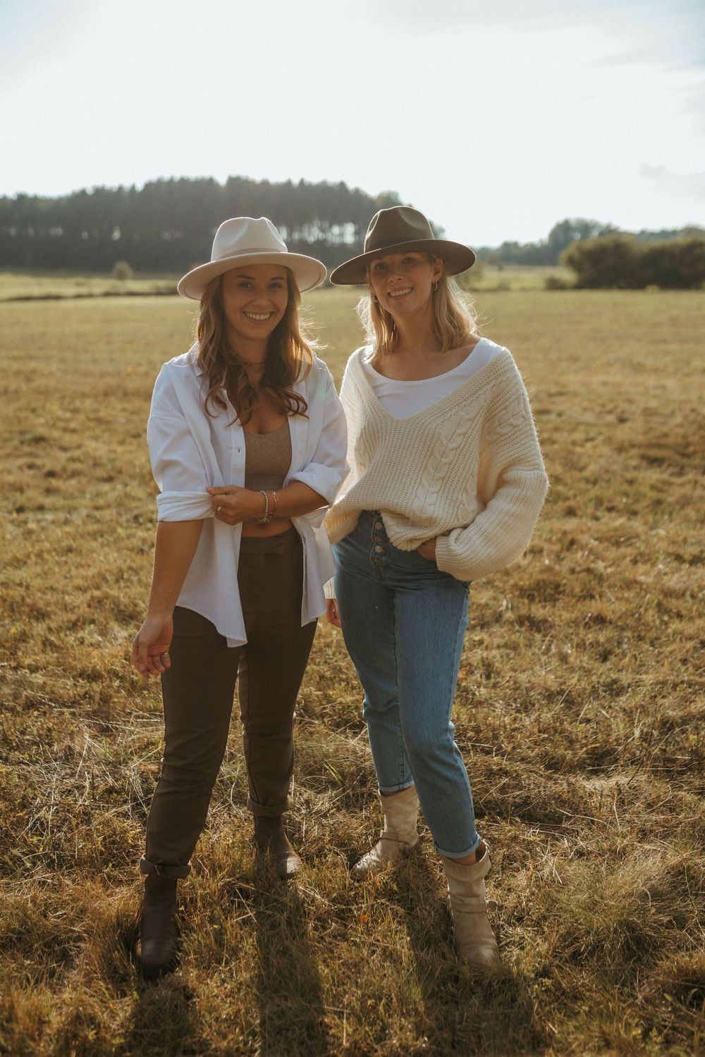 Two friends in hats and casual wear share joyful laughter while standing together in a golden autumn field at sunset.