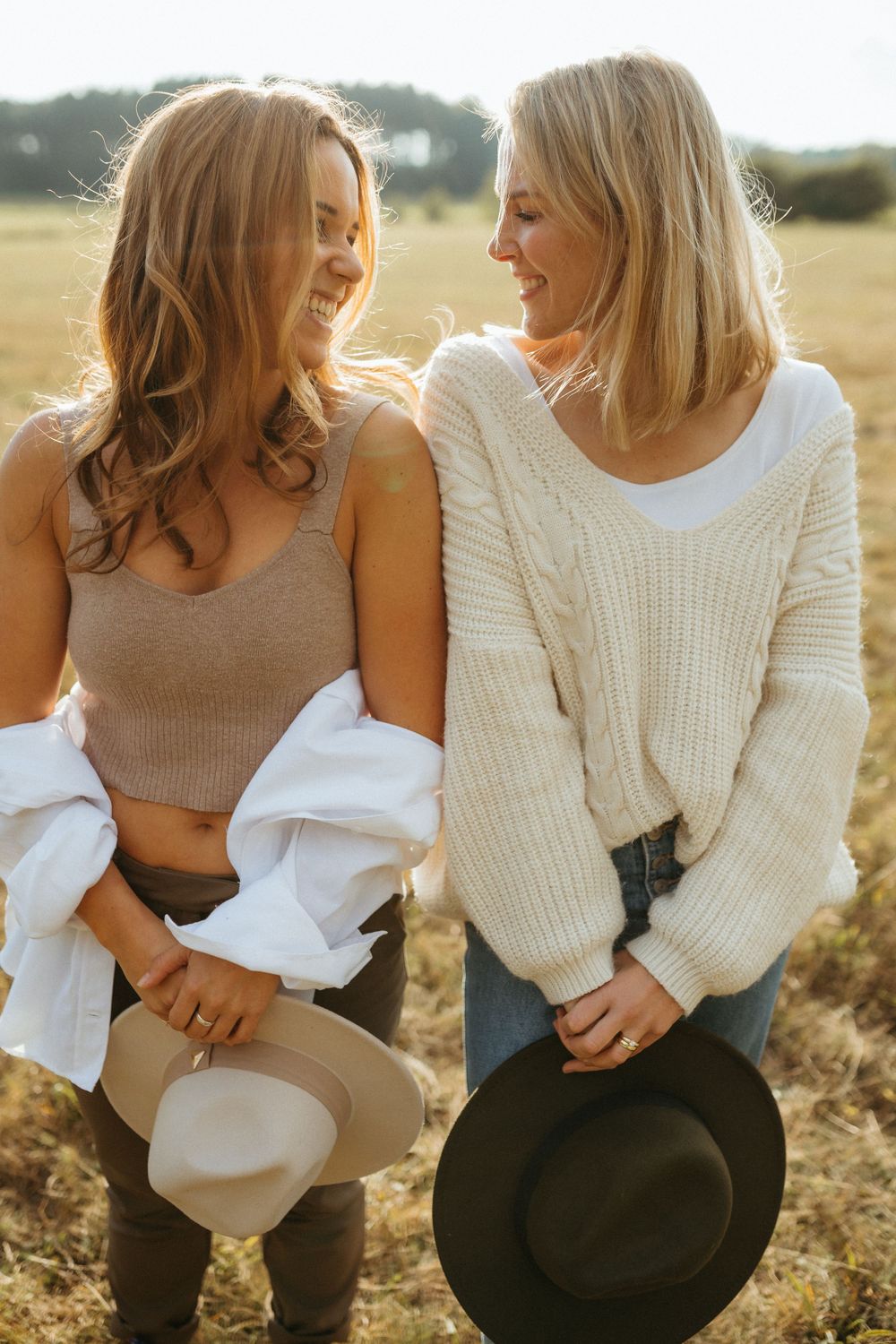 Two friends in cream and white sweaters sitting together in a sunlit field sharing joyful moments.