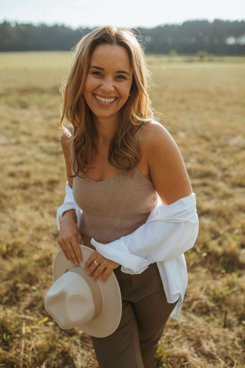 Casual portrait of someone holding a western hat while smiling brightly in warm sunset light.