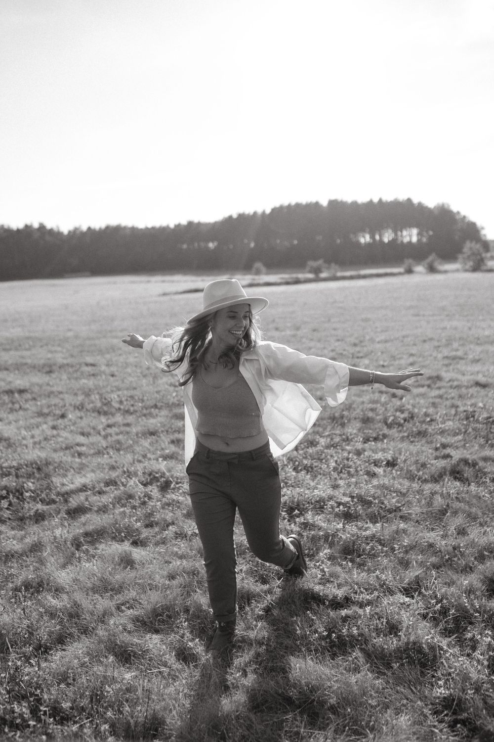 Two people in hats dance freely across a sunlit field with their arms outstretched.