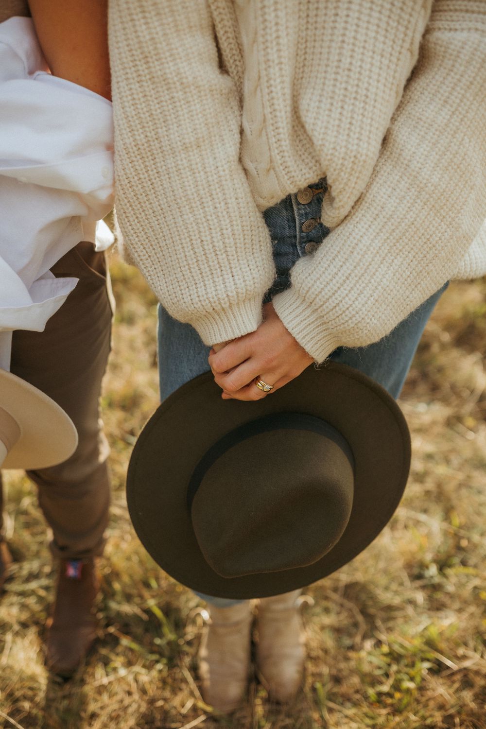 A person in a cream sweater holds a dark brown fedora hat against an autumn outdoor background.