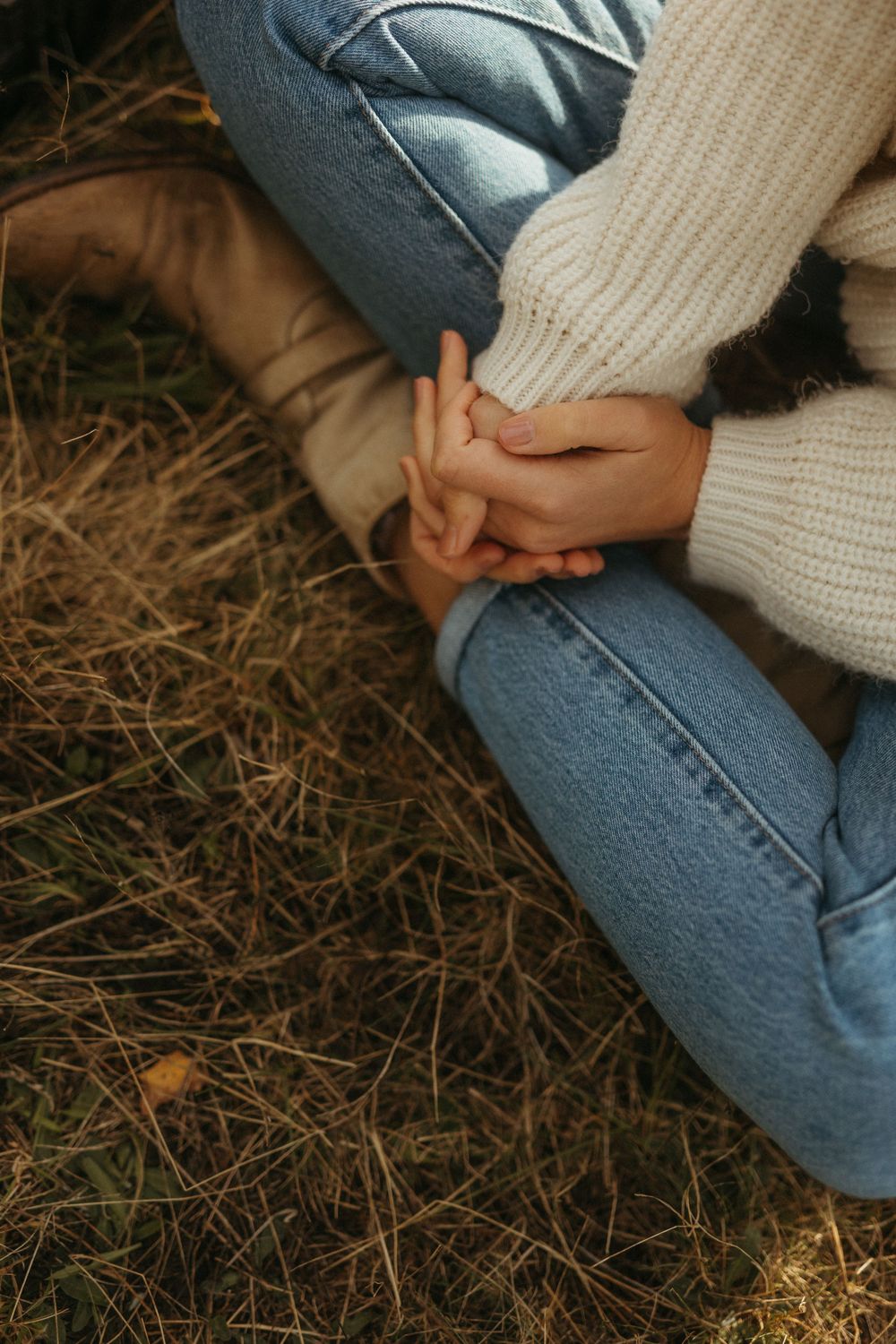 Person wearing blue jeans and white sweater sitting on dried grass in a natural setting.