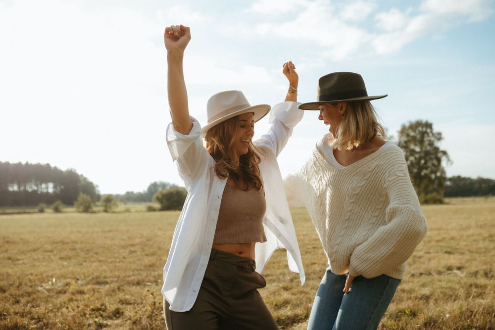 Two people dancing and celebrating in a sunny meadow wearing casual outfits and hats.