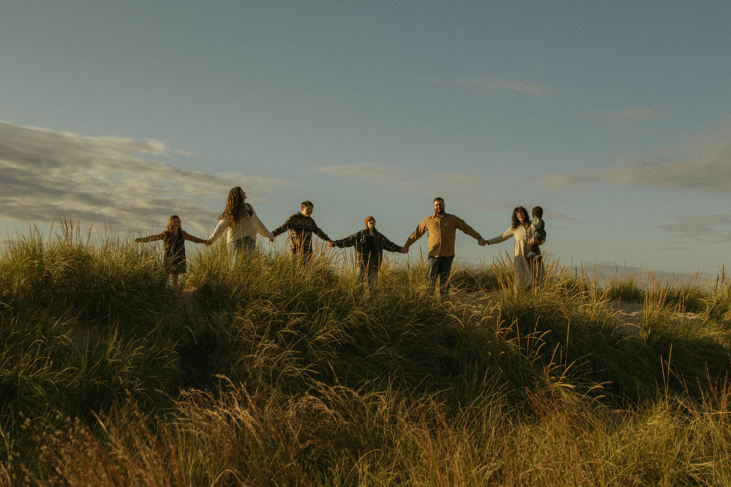 Group of people holding hands in a line on grassy coastal dune at sunset.