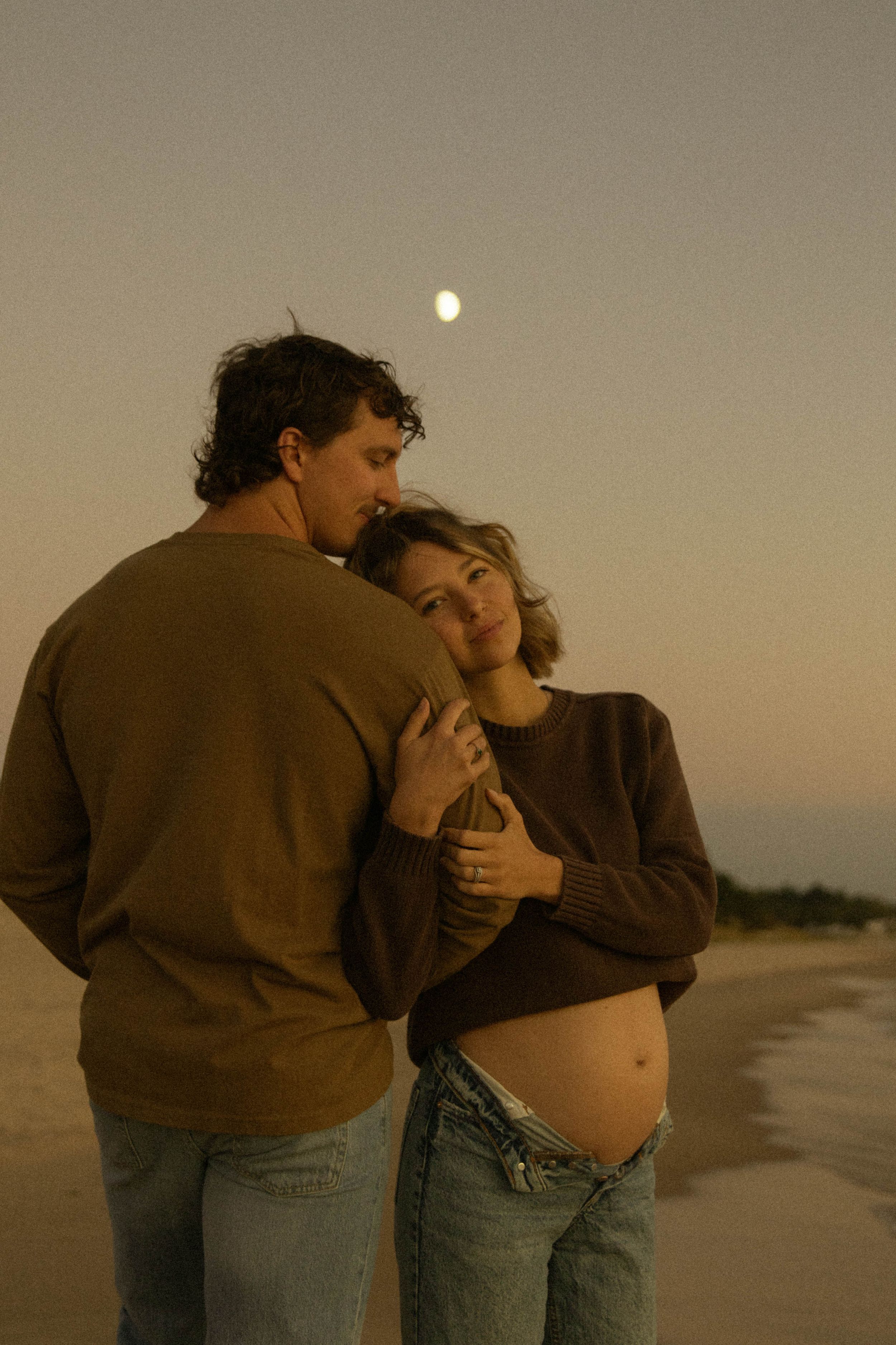 Couple embracing on beach at dusk with full moon, pregnant woman in brown sweater and jeans leaning into partner.