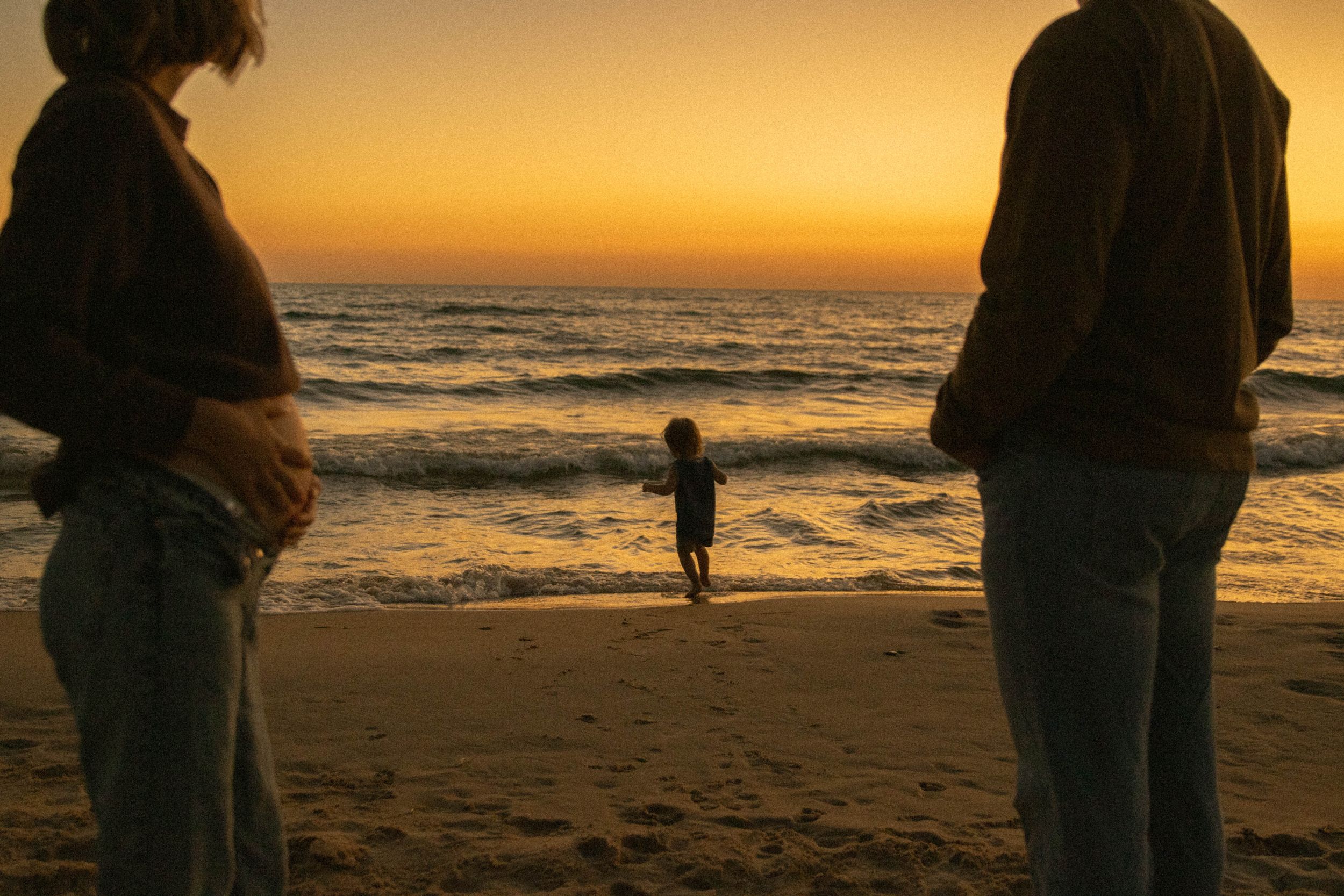 Family silhouetted at golden sunset beach, pregnant mother and partner watching child near ocean waves.