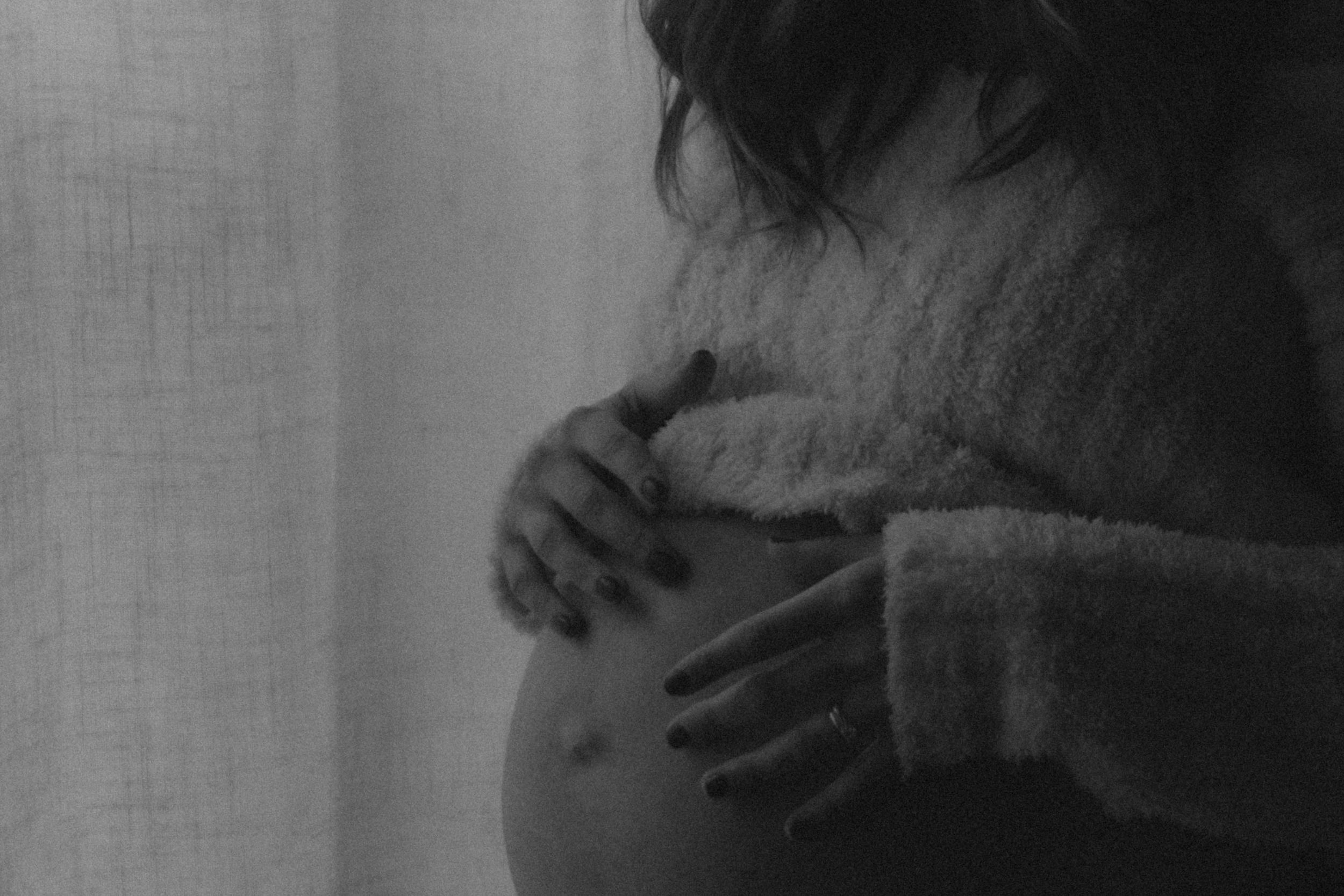 Black and white silhouette of pregnant woman gently holding baby bump near sheer curtain.