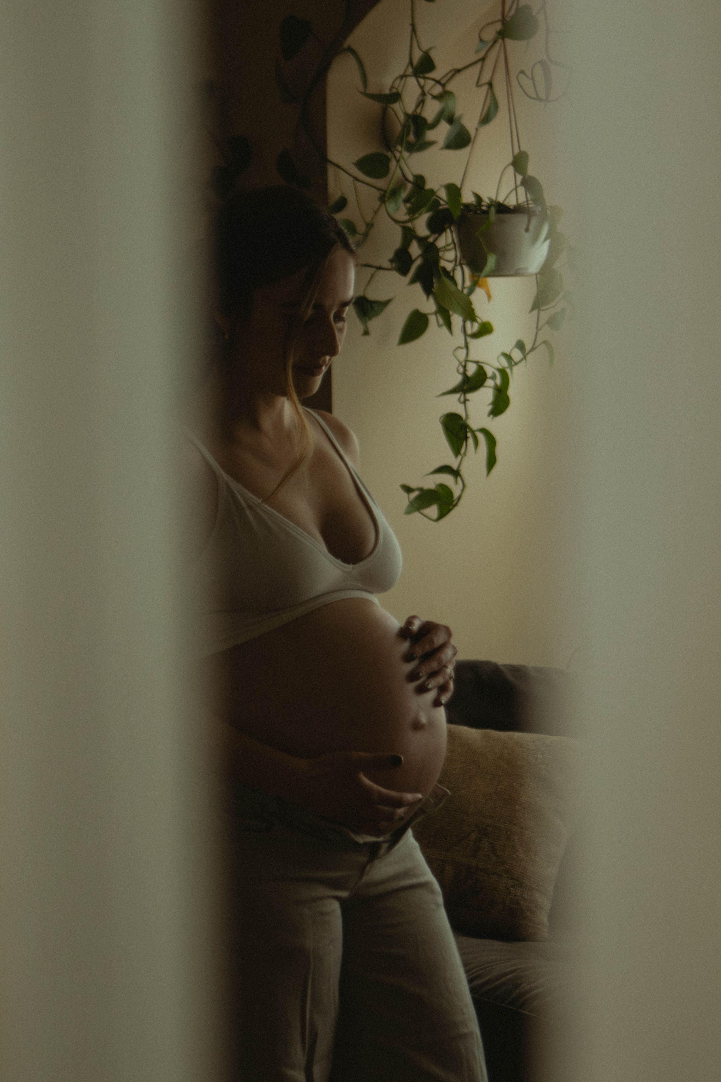 Pregnant woman in white crop top cradling her baby bump, standing by a window with hanging green plants.