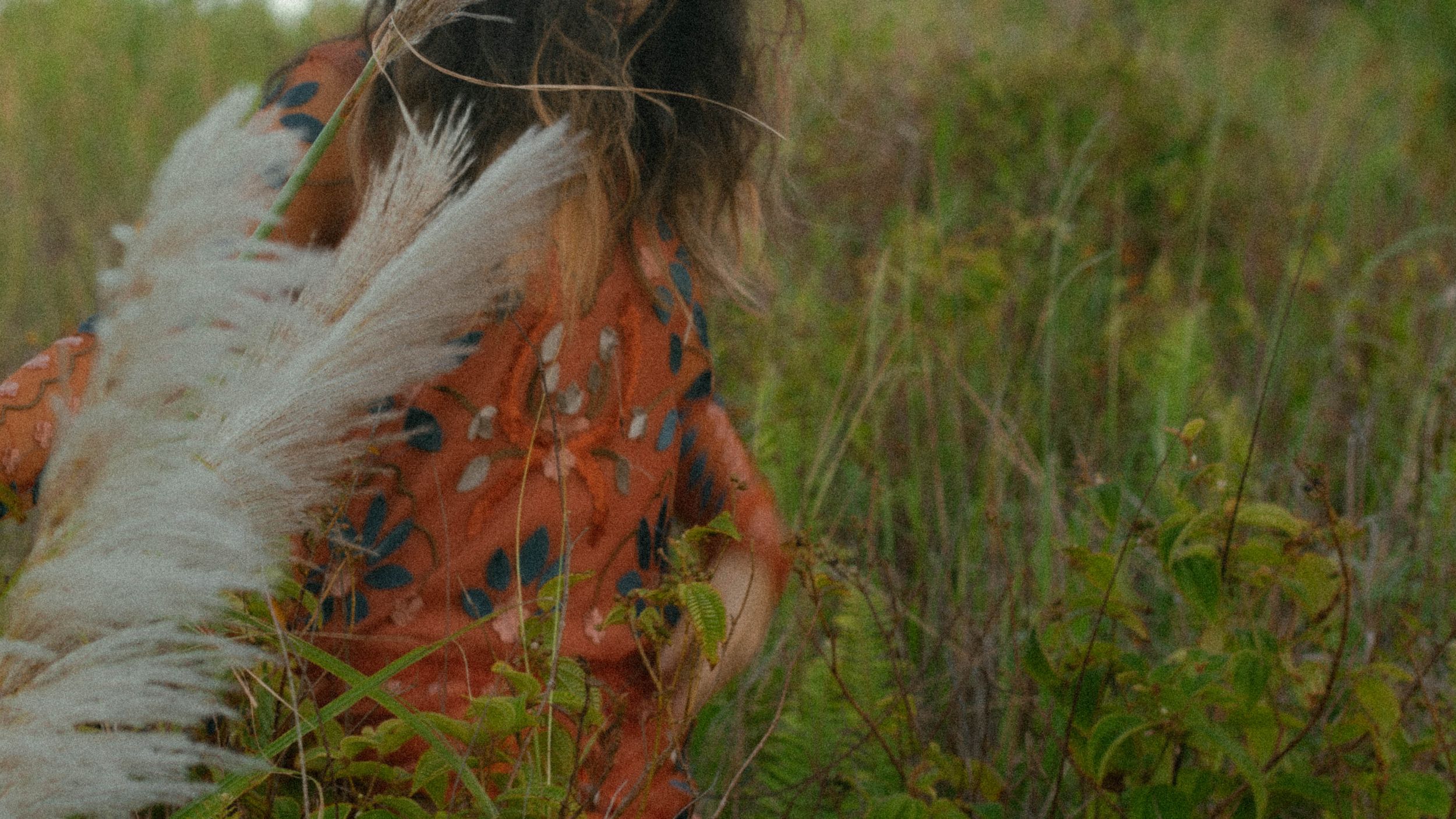 Woman in orange floral dress holding white pampas grass in a green field.