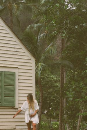 Pregnant woman in white dress standing beside a cottage surrounded by palm trees.