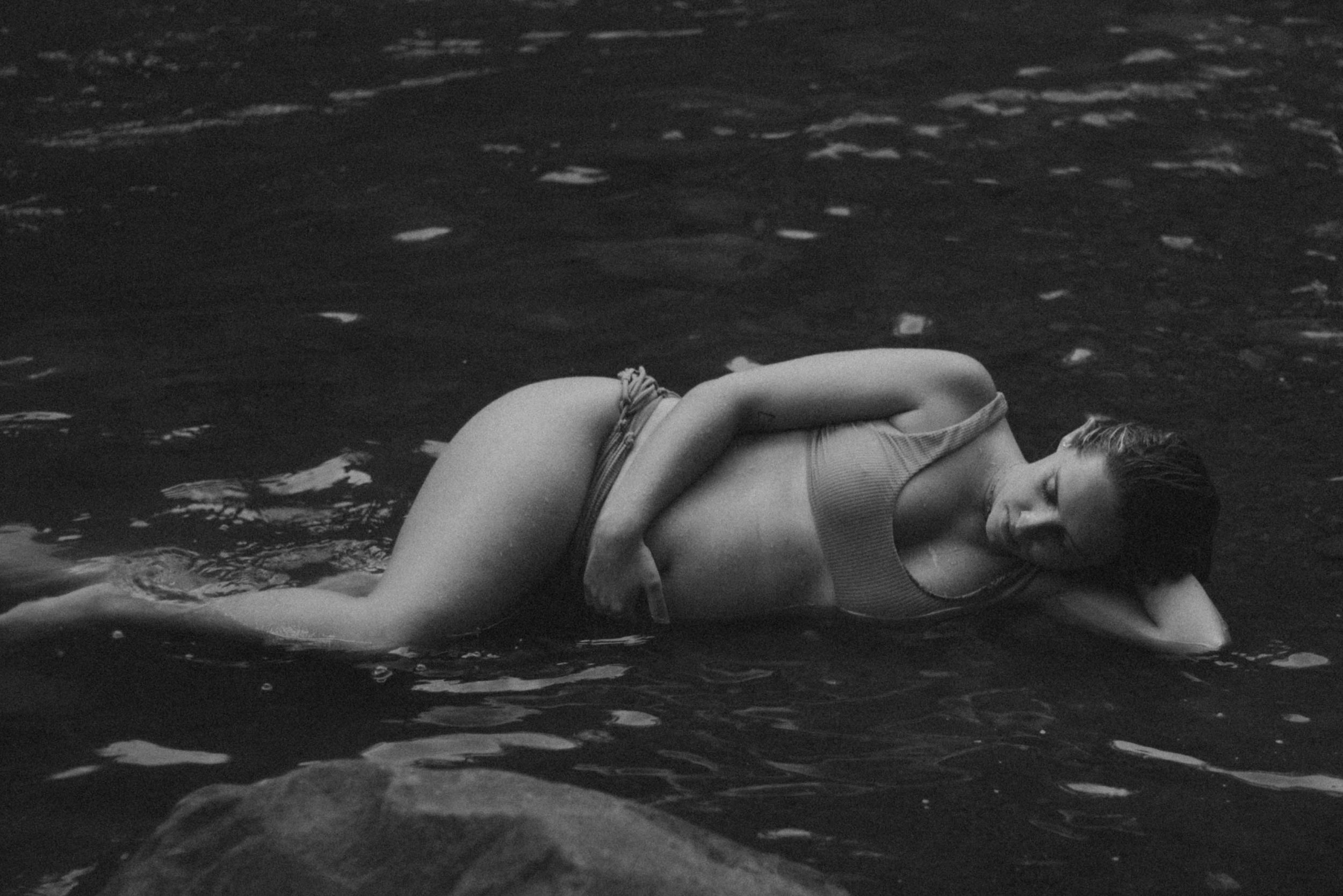 Black and white photo of a woman lying in shallow water on rocks.