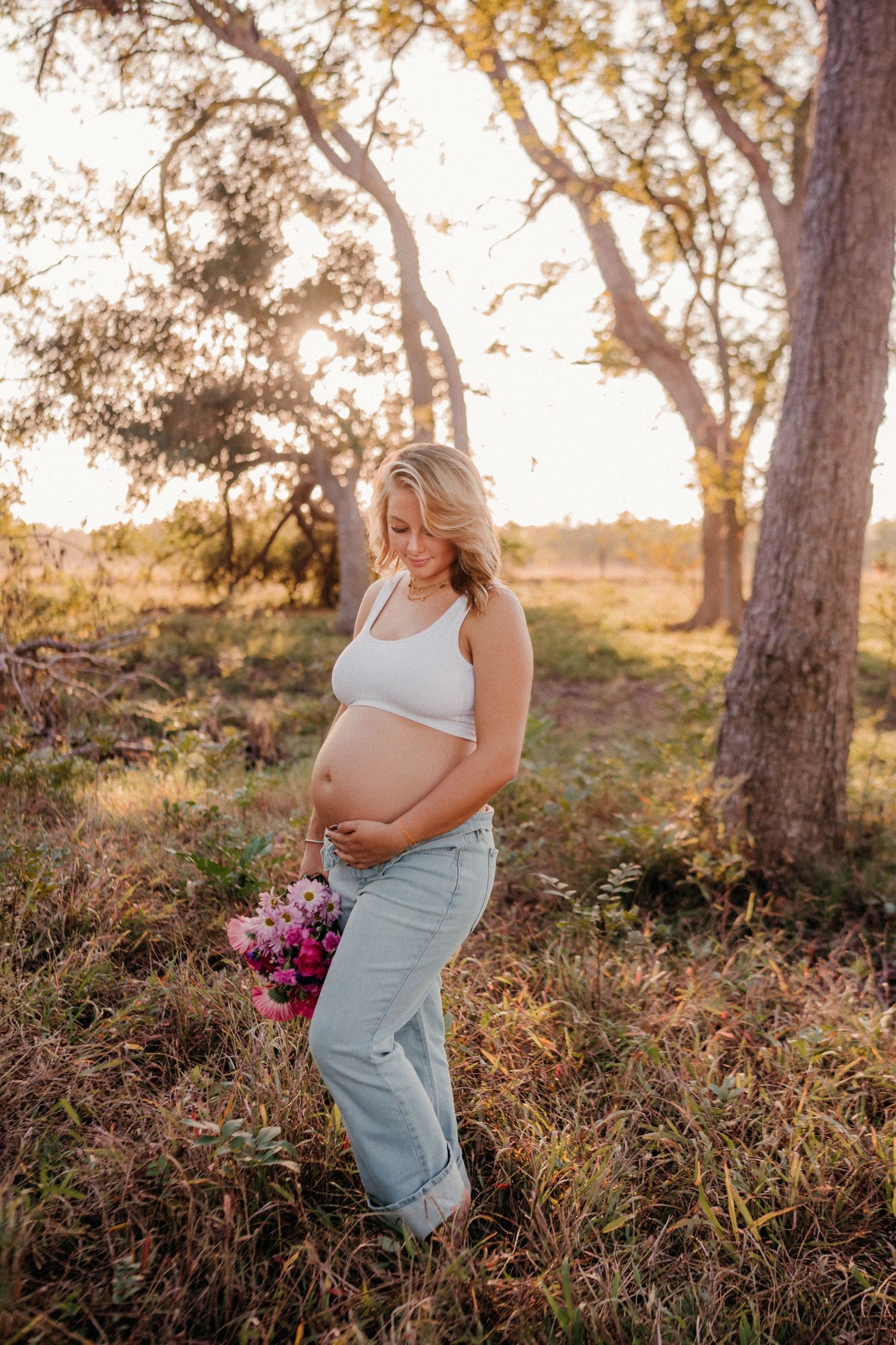 Expectant mother in white top and jeans holds purple flowers in golden sunset light among trees in nature setting.
