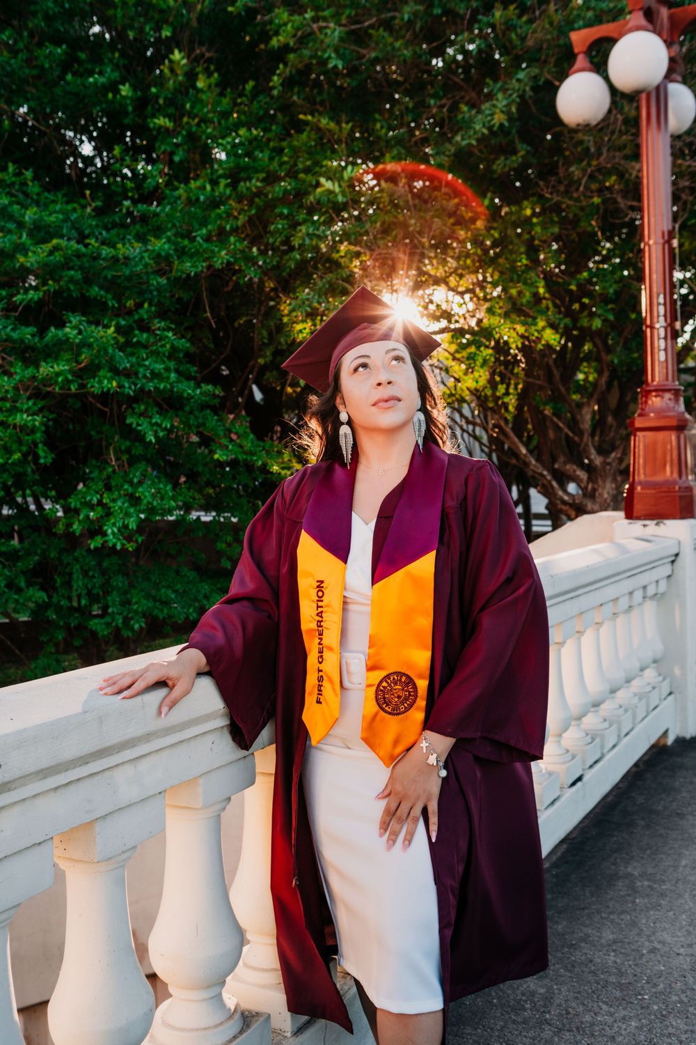 Graduate in maroon cap and gown with yellow stole poses on white balustrade during sunset at outdoor ceremony.