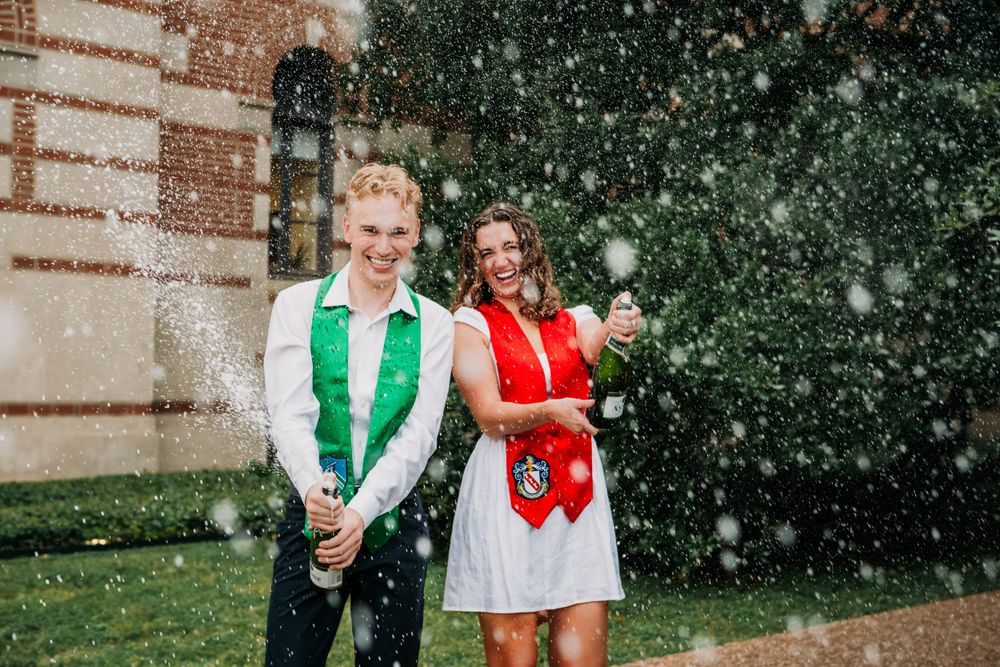 Two people in festive vests laugh and enjoy falling snow outside a brick building. Rice University