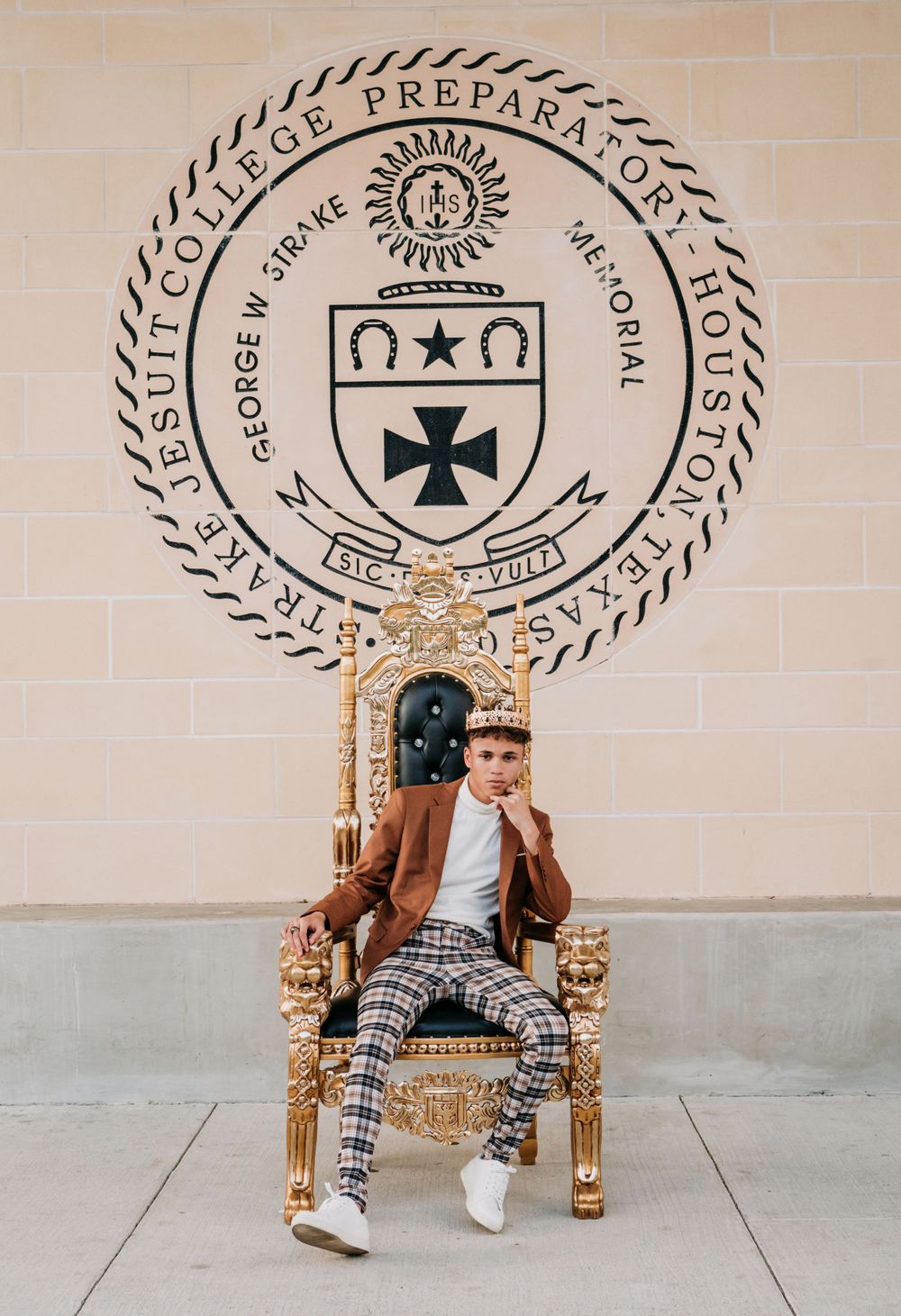 Ornate golden throne sits beneath a circular preparatory school crest and seal mounted on beige wall.