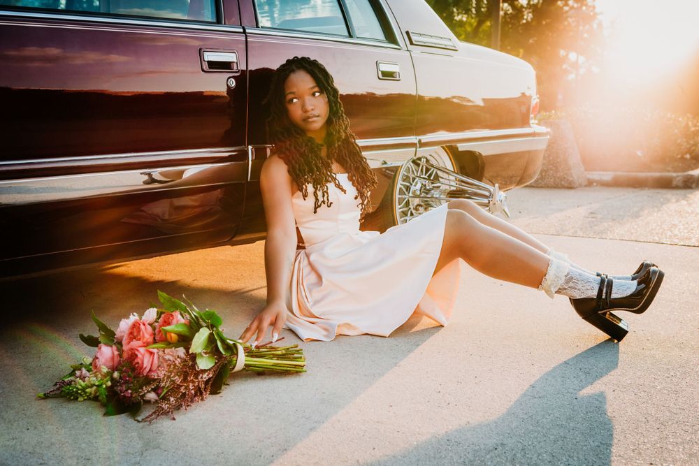 Someone in a white dress sits on pavement next to vintage car and flowers during sunset with dreamy lighting.