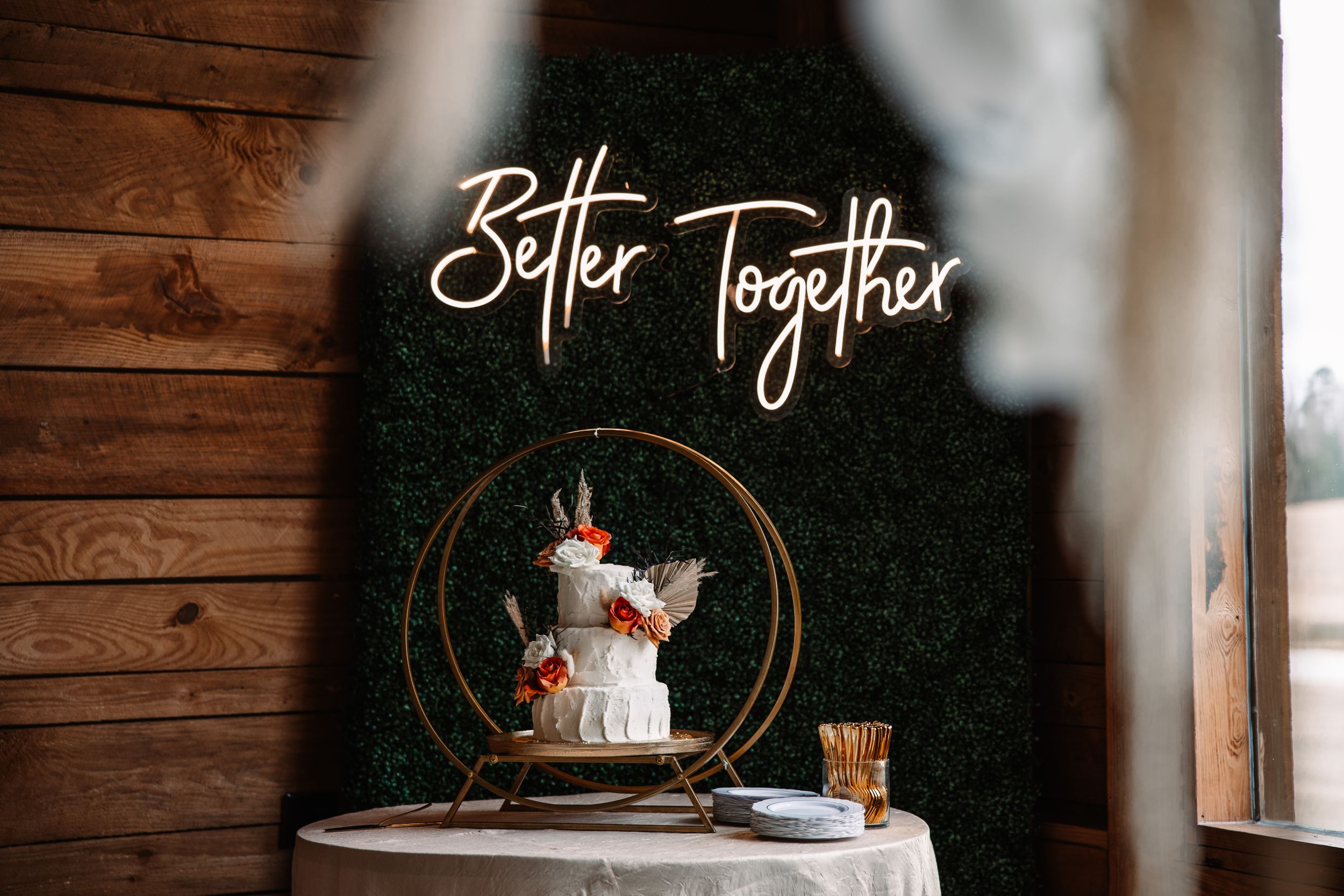 Wedding cake displayed under neon 'Better Together' sign with rustic wooden backdrop.